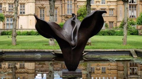 A brown, bronze sculpture called La Promesse, sitting in water in front of a grand building, which has intricate stonework, windows and columns. The artwork is based on the seed pod of the Iris Foetidissima plant.