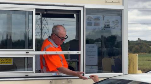 A man in a toll booth wearing a flourescent orange high-vis t-shirt collects a coin from the hand of a person stretching out of a car window.
