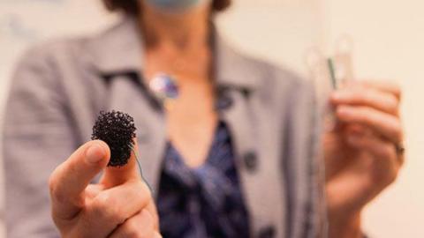 Prof Rebecca Fitzgerald in the background, wearing a mauve jacket over a purple top and with a surgical mask on. She is holding in the foreground a black sponge in her right hand which has a string attached