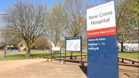 Outside entrance of New Cross Hospital, trees in the back ground with a sign with NEW CROSS HOSPITAL on it with writing saying emergency department below