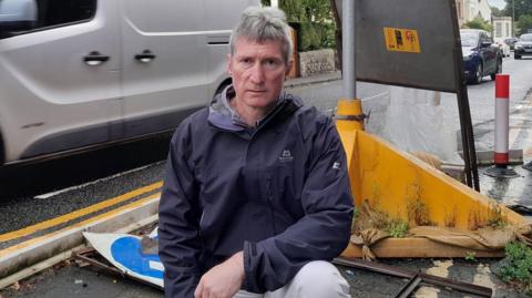 Peter Roberts looks into the camera with three vehicles travelling on a road behind him