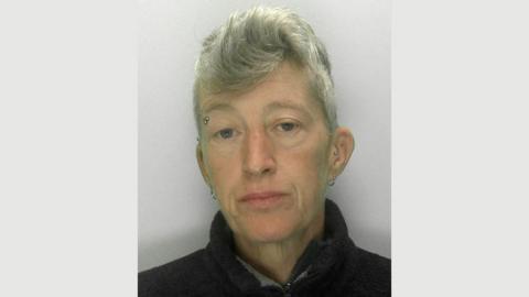 A police mugshot of Samantha Zala wearing a black pullover and standing against a grey background. She has short grey hair, an eyebrow piercing and two silver hoop earrings. 