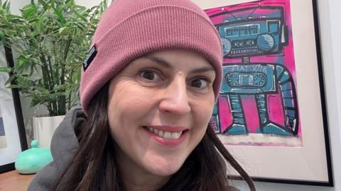 Sharon Davidson, woman wearing pink warm hat and smiling at camera, with a bright painting in the background.