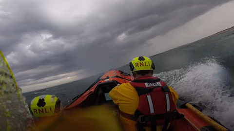 A view from behind of two RNLI crew members. They are on a lifeboat with waves splashing about. The sky above them is dramatic - dark and cloudy.