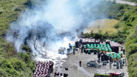 Scores of wheelie bins are seen from a distance. Some of them are on fire. 