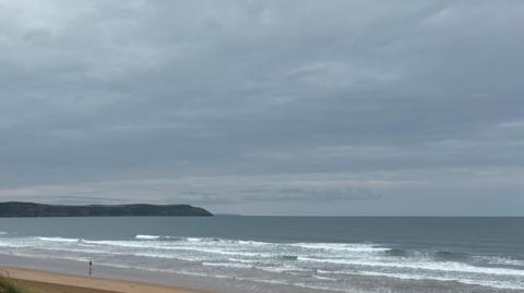 Woolacombe Beach in Devon. The beach is sandy and there are large waves in the distance. There is land on the left and people stood on the beach.
