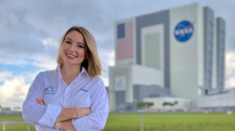 A picture of Sian Cleaver with her arms folded and posing in front of Nasa's large building at the Kennedy Space Centre in Florida. She has blonde hair and is wearing a white shirt. 