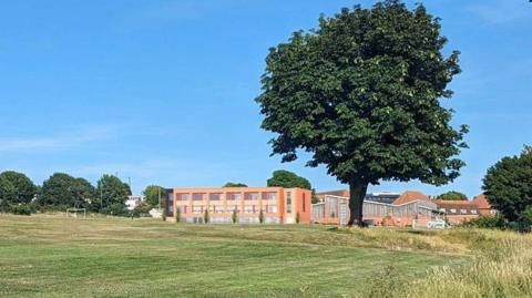 Parkland on a sunny day with blue skies and trees and an architect's image of the new three storey, red brick college building. 