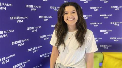 Pictured is Sarah O'Connor smiling against a purple background with the words BBC RADIO WM printed on it. She has brown hair with bleach blonde highlights. She is wearing a white t-shirt and blue jeans