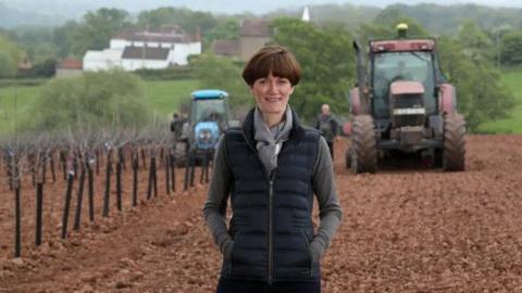 A woman wearing a grey long-sleeved top, grey scarf and black gilet with short brown hair stands in front of a field and two tractors. Countryside and buildings can be seen in the background.
