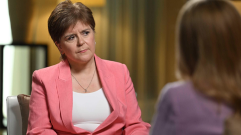 Sturgeon has short brown hair and is wearing a pink jacket and white ti-shirt as she is interviewed by a woman who has her back to the camera