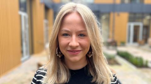 Sophie is standing in the courtyard of Shankill Shared Women's Centre. She has a blond middle parting with layers, gold large earrings on and is wearing a navy jumper with white stripes on it. 