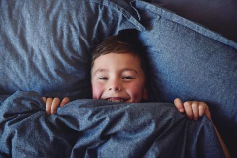 a boy smiling while lying on pillows pulling up a duvet up to his chin