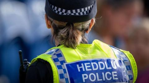 The back of a female police officer looking ahead of her on a street which is blurred. Her high-vis yellow police vest can be seen from behind. There is a large blue badge across the back which reads "Heddlu Police" in large white capitalised font. She wears a black hat and has her blonde short hair tied back in a low ponytail. 