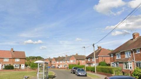 A residential street with houses, parked cars, a bus stop and grass verges.
