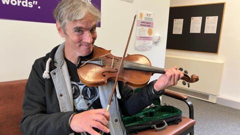 Man in a black hoodie and black T-shirt holds a violin next to a battered old violin case 