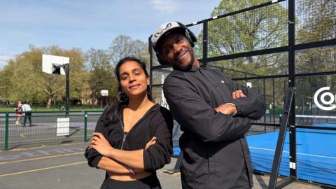A women with dark hair and wearing a black zip top and trousers stands next to a man wearing a baseball cap and black jacket. Behind them is a padel court and basketball court. They are both looking at the camera and smiling. 