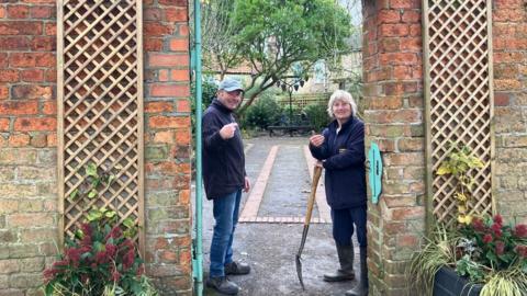 Volunteer Gary Bosworth in a navy fleece, jeans and boots and a cap, smiling and pointing at the garden at the entrance. Chair of trustees Lynn Ritson is holding a spade, also wearing a navy fleece, jeans and wellies and beckoning people to walk in. Two flower pots can be seen at either side of the brick entrance, with a pathway, trees and bushes in the background.