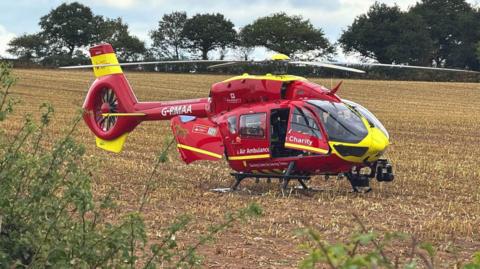 A red air ambulance that has landed in a field. The field has short yellow straw on the ground and a green bush is covering the
The helicopter's door is open. The nose of the aircraft is bright yellow as well as parts of the tail.