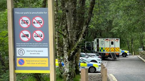Police cars and an ambulance parked in a car park in the background surrounded by trees. A sign in the foreground signals that the car park is unsuitable for certain large vehicles like trailers, lorries and buses 