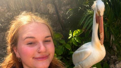Amy is smiling at the camera, she has ginger hair and brown eyes and nose piercings, she is wearing a blue t shirt. On her left is a large white bird with an orange beak.