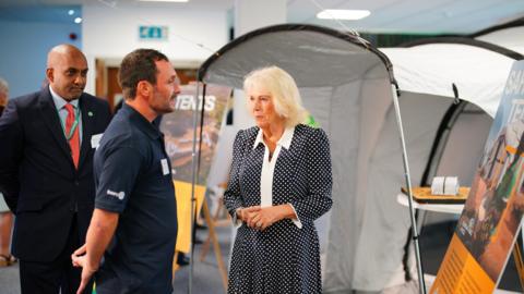 Queen Camilla is given a tour by the Chief Executive Officer of ShelterBox, Sanj Srikanthan who is standing to the left dressed in a suit, during a visit to the headquarters of the charity.
She is wearing a dark dress with white polka dots and a white collar which extends vertically down the front of the dress. She is talking to a volunteer dressed in a blue polo shirt and stands in front of a tent.