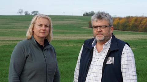 Rebecca and Philip Rayner are standing in their field, in the distance is a small hill called Chapel hill. Rebecca has a grey fleece on and black trousers, and Philip has a white and black checked shirt with a dark blue fleece. They have neutral expressions.