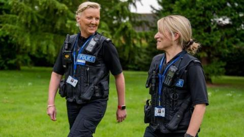 Two females in a park with trees and grass behind them. They are dressed in the navy blue uniform of special constables with the word POLICE on their left chest-pocket. Both are smiling and looking at each other. 