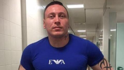 A man with short brown hair and a neck tattoo. He is wearing a dark blue top with white writing that reads 'EA' with a bird emblem. He also has a black tattoo on his upper arm.
