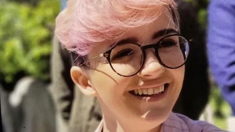 A woman with short pink hair, glasses and wearing a pink shirt.