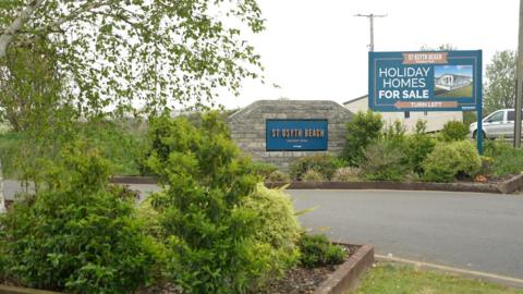 Blue signs at the entrance to St Osyth Holiday Park, which is based by the sea. A few static caravans are pictured in the background.