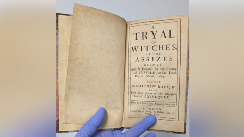 The historic book that recounts the Bury St Edmunds witch trials. It shows the first page of the book, which someone wearing blue gloves holds open.