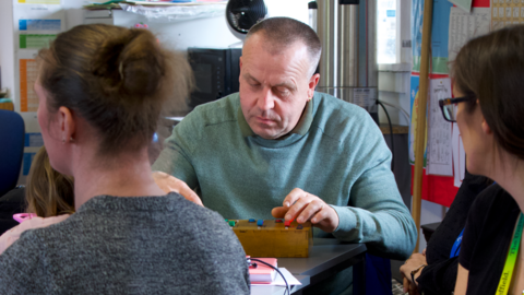 A man sat at a table during one of the University of Sheffield's dementia workshops, held in partnership with charity Age UK. He is sat at a table, focused on a wooden box with audio faders built into it. He's being watched by a woman wearing a university lanyard.