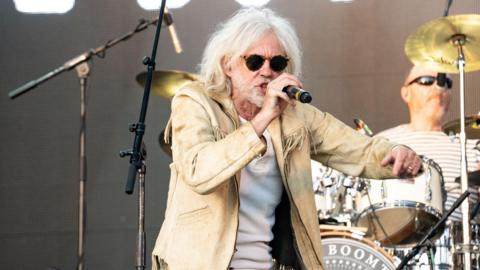 Bob Geldof of The Boomtown Rats singing with his band onstage, wearing a beige suede jacket with tassels