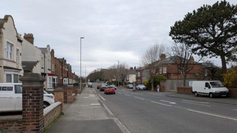 Hartburn Lane which has houses on either side of a road with cars driving on both sides. The homes in the foreground are white with driveways. The houses further back are red brick. There is a bus stop on the right side of the road and a van parked on the path in front of it. The sky is grey.