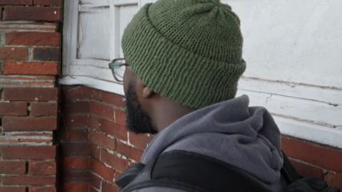 A man wearing a green hat and wearing a grey hoodie and a black rucksack sits facing away from the camera next to a white and brick wall. He has a beard and is wearing glasses.