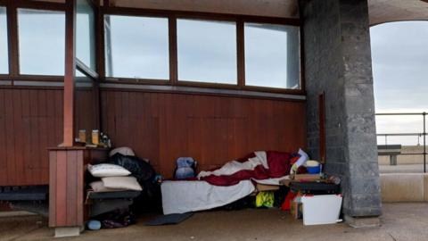 A person lying down huddled in a blanket in a seaside shelter. It has wooden panels and windows