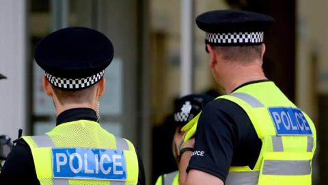 Two police officers stood talking to a third while dressed in police uniform, including black and white hats, high-vis vests and walkie talkies. 