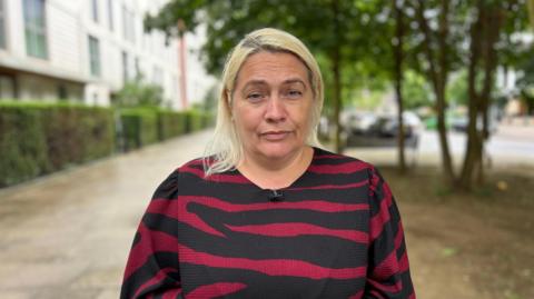 A woman with light blonde hair standing outdoors on a residential street, wearing a black and red striped top, with blurred buildings, trees, and parked cars in the background