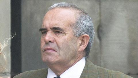 A man with short grey hair and black eyebrows pictured outside. He is wearing a green checked jacket and a shirt and tie