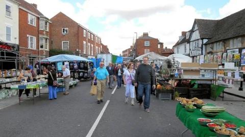 Archive photograph of Grantham market. There are a number of stalls covered in tarpaulins and people walking along looking at the goods that are for sale.