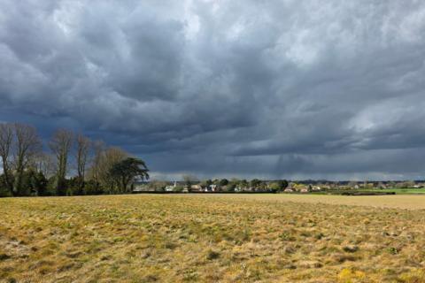 Dark storm clouds over a rural landscape, with rain falling in the distance beyond a grassy field and a line of trees.