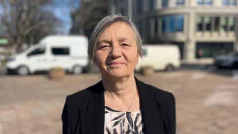 Deputy Mary Le Hegarat is standing in a paved square in front of a road, with an office building behind it. She is wearing a black blazer and is looking at the camera.