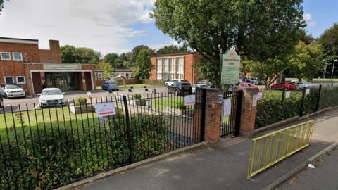 Google Streetview image of front of Leagrave Primary School showing public pavement, school sign, fencing and hedging, car park and front of school building