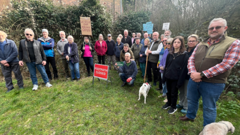 A group of people of all ages are gathered together with signs saying things like 'we need our bridge open'. Some people have their dogs with them. They are stood behind a red 'footpath closed' sign. They're stood outside with trees behind them. 