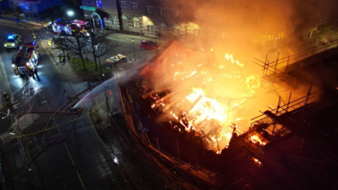 An aerial view of a building well alight with several fire engines parked up and firefighters spraying water on to the flames.