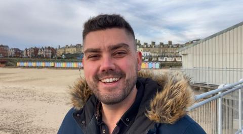 A man with short dark hair and a beard smiles at the camera while standing on a beach on a grey day. He is wearing a navy coat with a faux fur lined hood. He has a black shirt on underneath. Beach huts on the beach can be seen in the distance as well as some homes. 