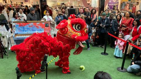 A red and gold lion costume which is 'eating' a lettuce. Around him are people watching and taking photographs. 