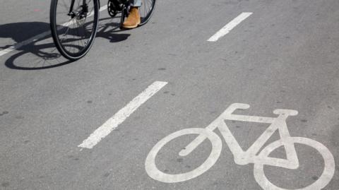 A cycle lane with a bike painted on the floor in white paint with the wheels and pedals of a bike seen on the other side of the lane.