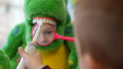 Child brushing a dinosaur's teeth while holding a magnifying glass over the teeth